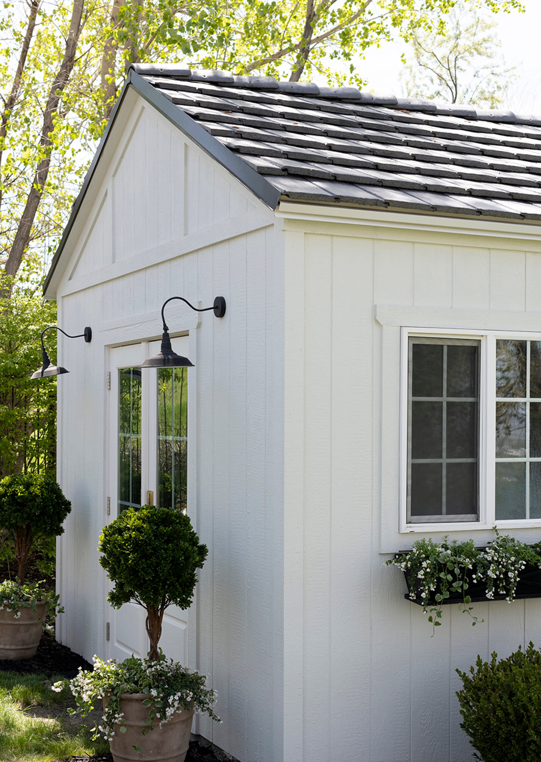 Boxwood topiaries planted in planters with trailing flowers, flanking french doors on a storage shed. Window flower boxes are also installed above a row of boxwoods on the opposing side of the shed.