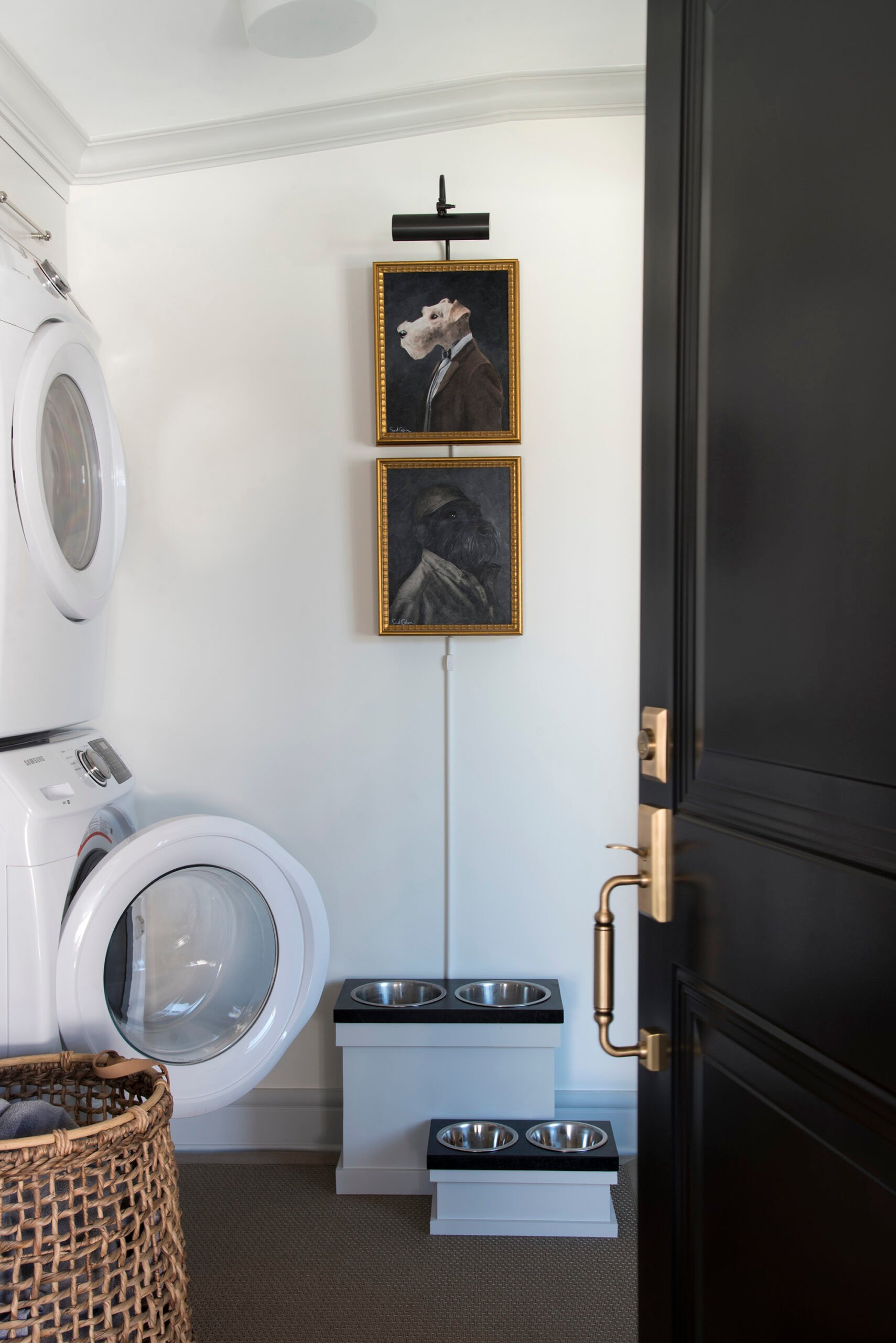 Laundry room with stacked washer and dryer, stacked dog portraits beneath a picture light with a hidden cord, custom soapstone dog feeders, and a woven rug, and hamper with a black door opening into the room